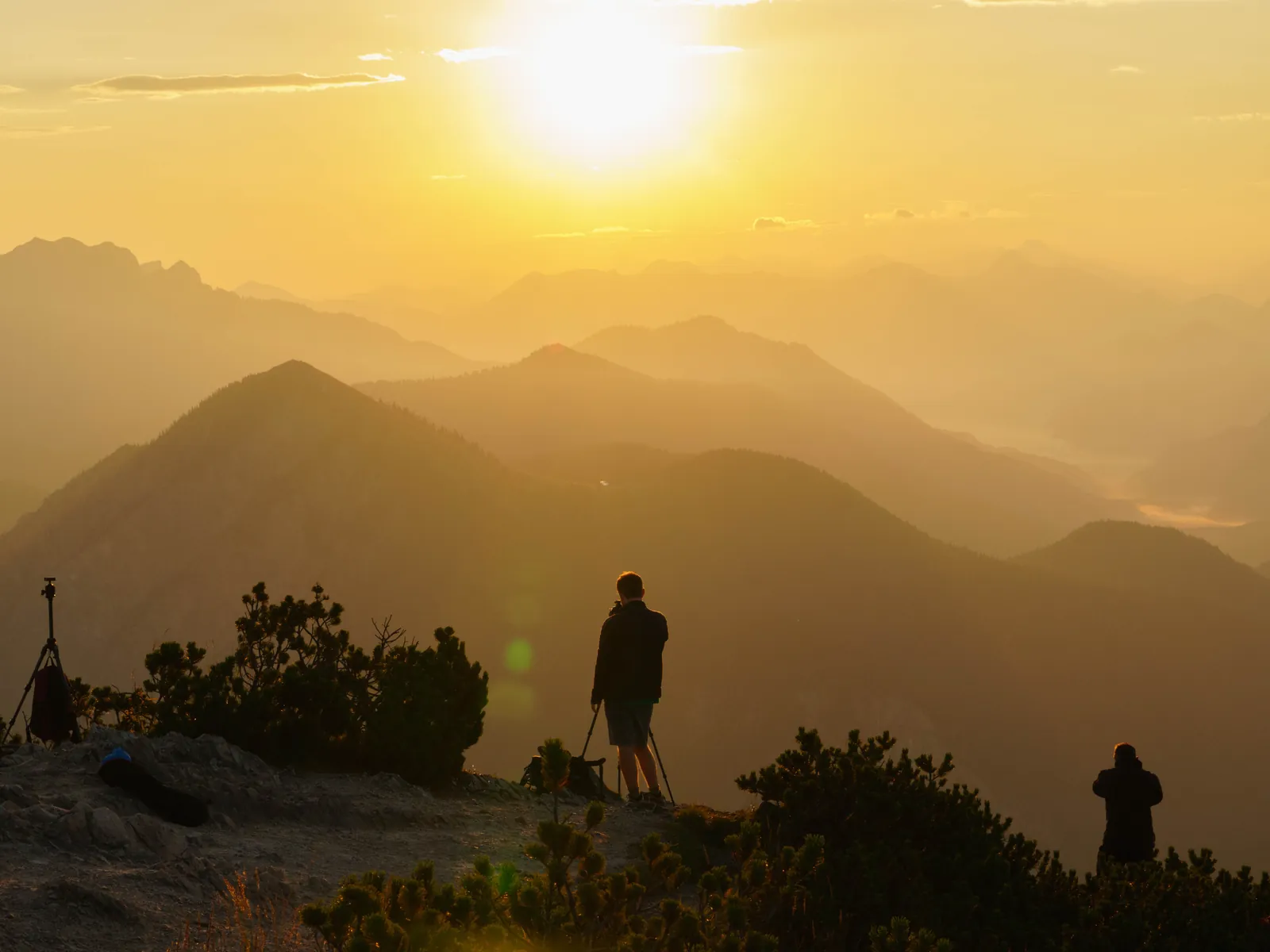 A photographer with a tripod and backpack on a summit, backlit by the evening sun